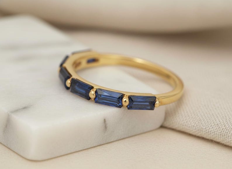 May include: A gold ring with a row of rectangular, deep blue gemstones. The ring is set on a white marble surface, with a soft, neutral-colored fabric in the background. The ring's band is a polished gold.
