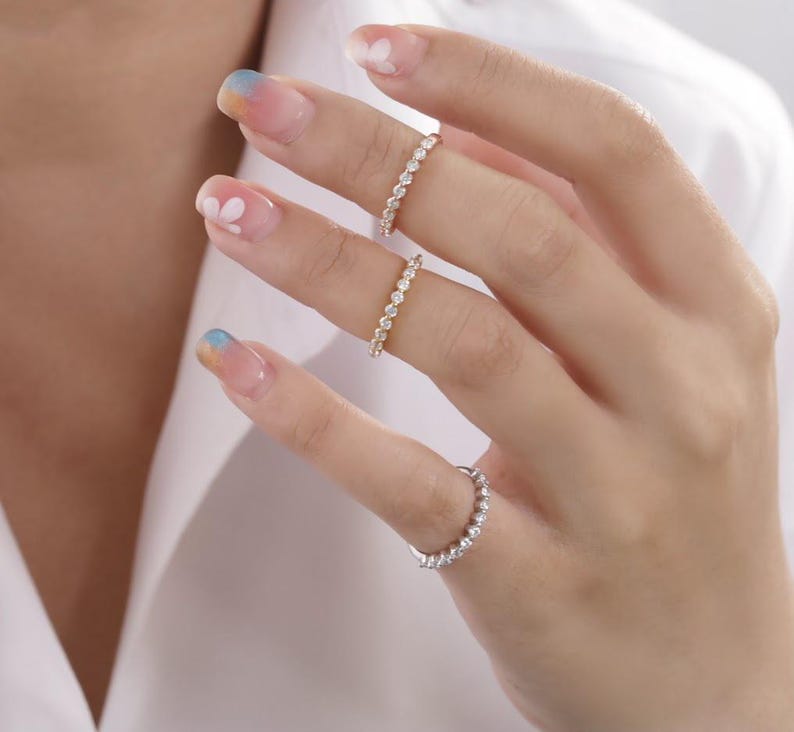 May include: Three delicate gold rings with small clear stones set in a continuous line. The rings are worn on the fingers of a hand with painted nails.