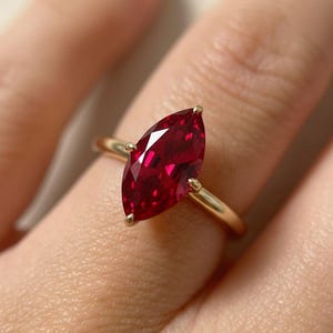 May include: A gold ring featuring a marquise-cut, deep red gemstone. The gemstone is held in place by four prongs. The ring's band is a simple, polished gold. The ring is shown on a finger.