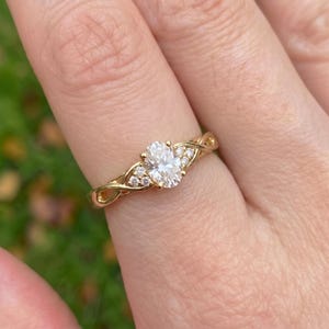Peut inclure: Une bague en or avec une pierre centrale transparente taille ovale, entourée de trois petites pierres rondes transparentes de chaque côté. L'anneau présente un design métallique torsadé et entrelacé qui soutient le sertissage.