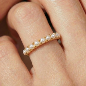 Peut inclure: Une bague en or ornée d'une rangée de petites perles blanches rondes. La bague est portée au doigt, mettant en valeur le design délicat et le contraste entre l'or et les perles. La bague est un bijou.