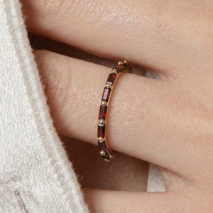 May include: A gold ring with a band set with rectangular, deep red gemstones and small gold beads. The ring is worn on a finger, showing its design and colour against the skin and a white fabric background.