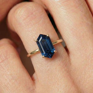May include: A gold ring featuring a large, rectangular, deep blue gemstone. The gemstone is the focal point, set with four prongs on a delicate gold band. The ring is shown on a person's finger.