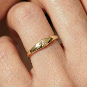 Puede incluir: Un anillo de oro con una banda lisa y pulida que se estrecha hacia el centro, donde una pequeña piedra preciosa transparente de corte redondo está engastada en un grabado en forma de estrella. El metal tiene un acabado cálido y brillante.