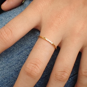 May include: A thin gold band ring featuring a row of five small, clear, round-cut stones set along the top. The metal has a polished, reflective finish, and the stones are arranged in a straight line across the center of the band.