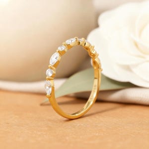 以下が含まれることがあります： 金色のバンドリング。マーキスとラウンドカットの透明な宝石が交互に並んでいます。リングは、白い花と緑の葉が見える、柔らかくニュートラルな背景に配置されています。