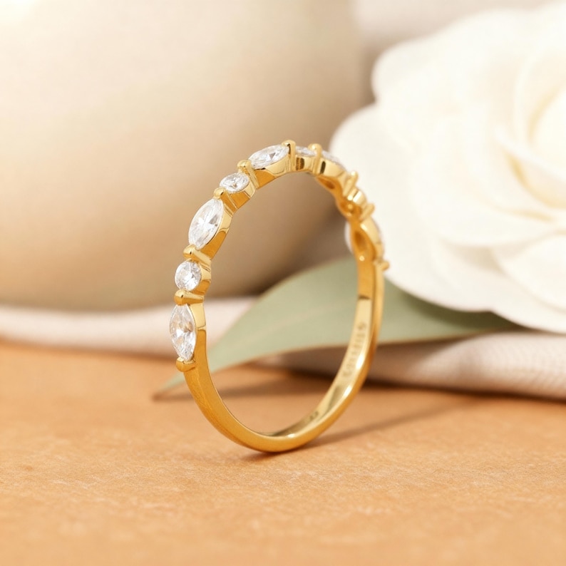 以下が含まれることがあります： 金色のバンドリング。マーキスとラウンドカットの透明な宝石が交互に並んでいます。リングは、白い花と緑の葉が見える、柔らかくニュートラルな背景に配置されています。