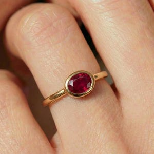 May include: A gold ring featuring a single oval-cut deep red ruby gemstone set in a smooth bezel setting. The band is crafted from polished yellow gold, creating a clean and simple look that highlights the vibrant color of the central stone.
