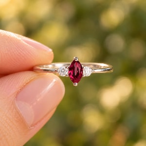 May include: A silver-toned ring featuring a central marquise-cut deep red gemstone flanked by two small, round, clear white stones. The band is thin and polished, showcasing a classic three-stone design with a vibrant, faceted center stone.