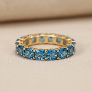 May include: A gold-colored ring featuring a continuous band of square-cut, light blue gemstones. The ring's design showcases a classic eternity style, with each gemstone set closely together, creating a seamless appearance. The ring is displayed on a neutral, textured surface.