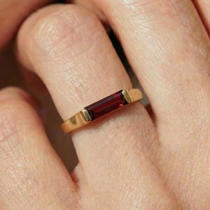 Puede incluir: Un anillo de oro con una gema rectangular de color rojo intenso. El anillo tiene una banda de oro pulido y simple, con la gema colocada horizontalmente. El color de la gema contrasta con el oro.