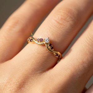 May include: A gold-colored ring with a wave design. The ring features a central, clear gemstone and several smaller, red gemstones. The ring is on a finger, with the focus on the jewelry.