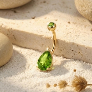 Puede incluir: Un piercing para el ombligo dorado con una piedra preciosa verde claro en forma de pera. La barra curva tiene una pequeña gema verde redonda en la parte superior. La joya está colocada sobre un fondo neutro con textura arenosa.