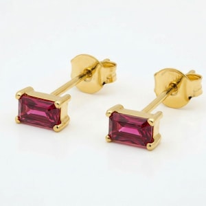 May include: A pair of gold stud earrings, each with a rectangular, deep pink gemstone. The earrings have a classic design with a post and butterfly backing. The gemstones are set in a gold bezel.