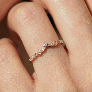 Può includere: Un anello in oro rosa caratterizzato da una delicata fascia a forma di V con un motivo a foglie testurizzate. La fascia è impreziosita da piccole pietre chiare a taglio rotondo incastonate lungo il fogliame metallico, creando un design organico.