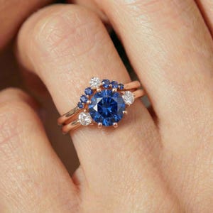 May include: A rose gold ring set featuring a large, round, deep blue gemstone as the focal point. The ring is accented with smaller white and blue gemstones. The band is a delicate rose gold color.