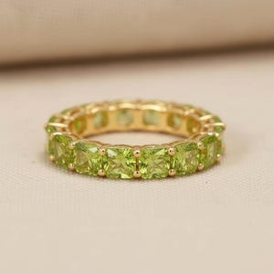 May include: A gold ring with a continuous band of square-cut, light green gemstones. The ring is set on a neutral-colored surface, showcasing the vibrant green stones and the warm gold setting.