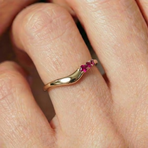 Könnte beinhalten: Ein goldener Ring mit einer glatten, geschwungenen Ringschiene, die spitz zuläuft. Der Ring ist mit drei kleinen, runden, leuchtend rosaroten Edelsteinen besetzt, die in einer Reihe entlang der oberen Kurve des Rings angeordnet sind.