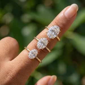 May include: Three gold rings featuring oval-cut clear stones set in four-prong settings. The rings are arranged in a vertical row, each showcasing a polished gold band and a brilliant, faceted center stone that reflects light with clarity.