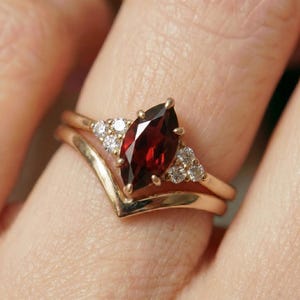 May include: A gold ring set featuring a marquise-cut, deep red gemstone as the centerpiece, flanked by smaller, clear gemstones. The ring is paired with a chevron-shaped band, creating a layered effect. The setting is a warm gold color.