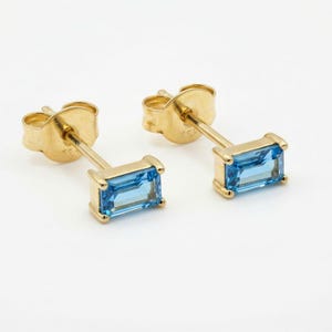 May include: A pair of gold stud earrings, each featuring a rectangular, light blue gemstone. The earrings have a simple, elegant design with a post and backing for secure wear. The gemstones are set in a gold bezel.