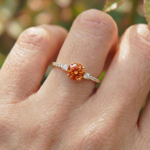 Op de afbeelding: Een gouden ring met een ronde, oranje edelsteen als middelpunt, geflankeerd door kleinere, heldere stenen. De ring zit om een vinger, met een vage achtergrond van groen gebladerte.