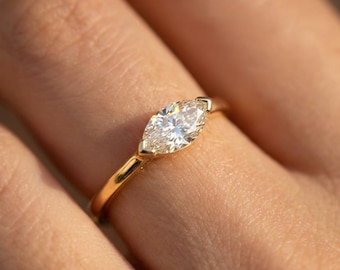 May include: A gold ring featuring a marquise-cut diamond. The diamond is set in a four-prong setting, and the band is a simple, polished gold. The ring is shown on a finger, with the focus on the diamond and the band.