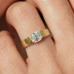 Puede incluir: Un anillo de oro con un diamante de talla cuadrada. El diamante está engastado en un engaste de canal, con la banda de oro que se extiende a ambos lados de la piedra. El anillo es un diseño clásico, adecuado para diversas ocasiones.