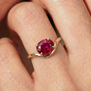 Op de afbeelding: Een gouden ring met een enkele ovaal geslepen dieprode edelsteen vastgehouden door vier pootjes. De band heeft een glad, gebogen, golfachtig ontwerp dat taps toeloopt naar de zetting, wat zorgt voor een vloeiende uitstraling.