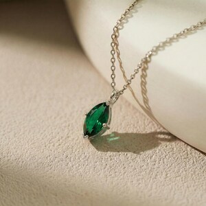 May include: A silver necklace featuring a marquise-cut, emerald-green gemstone pendant. The pendant is set in a silver bezel and hangs from a delicate silver chain. The necklace is displayed on a textured, neutral-toned surface.