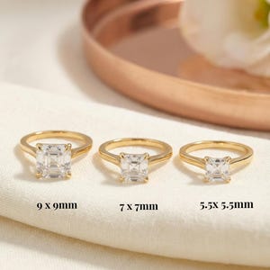 May include: Three gold engagement rings with square-cut center stones are displayed on a white surface. The rings are labeled with their dimensions: 9 x 9mm, 7 x 7mm, and 5.5 x 5.5mm. The rings are set against a soft, neutral background.