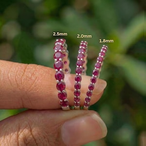 May include: Three silver rings with a continuous row of round, deep red gemstones. The rings are labeled with sizes: 2.5mm, 2.0mm, and 1.8mm. The rings are held against a blurred green background.