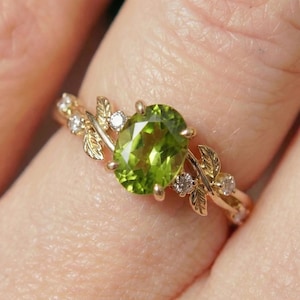 May include: A gold ring featuring an oval, light green gemstone. The ring is adorned with small diamonds and leaf-shaped accents. The ring is on a finger.