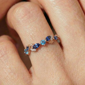May include: A rose gold ring featuring a leaf design with alternating blue and light blue gemstones. The ring has a delicate, nature-inspired aesthetic, with small white accent stones.