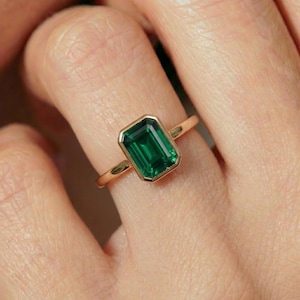May include: A gold ring featuring a rectangular, emerald-cut green gemstone. The gemstone is set in a gold bezel setting, and the band is a simple, polished gold. The ring is shown on a person's finger.