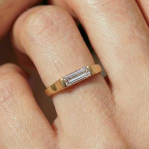 Può includere: Un anello d'oro con una pietra preziosa rettangolare e trasparente incastonata orizzontalmente. La fascia dell'anello è in oro lucido. La pietra preziosa è trasparente e riflette la luce. L'anello è un gioiello.