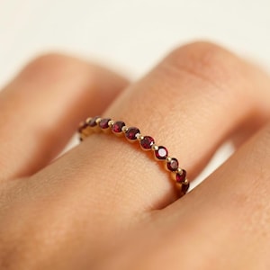 May include: A gold ring featuring a series of small, round, red gemstones. The gemstones are set closely together, forming a continuous line around the ring. The ring is displayed on a finger, showcasing its delicate design.