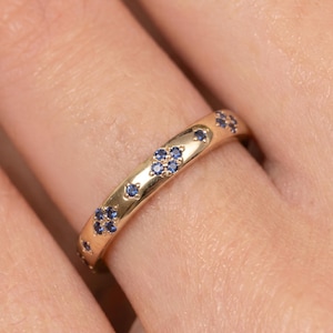 May include: A gold band ring featuring multiple small, round, blue gemstones set in clusters of four, arranged in a repeating pattern along the outer surface of the polished metal band, creating a distinct star-like or floral visual effect.