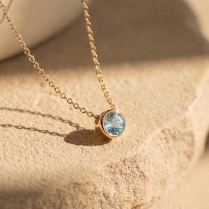 May include: A gold necklace featuring a round, pale blue gemstone pendant. The fine chain leads to a gold bezel setting holding the stone. The necklace is presented on a textured, light brown surface.