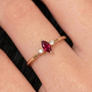 Op de afbeelding: Een gouden ring met een centrale robijn in marquise-slijpvorm, geflankeerd door twee kleine ronde witte diamanten. De ringband is dun en gepolijst, met een verfijnd ontwerp dat de levendige rode steen en de heldere accentstenen benadrukt.