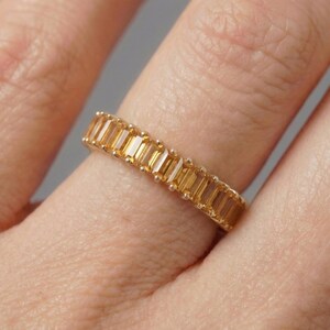 Könnte beinhalten: Ein goldener Ring mit einem Band aus rechteckigen, bernsteinfarbenen Edelsteinen. Die Edelsteine sind eng aneinander gesetzt und bilden eine durchgehende Farblinie. Der Ring befindet sich an einem Finger und zeigt das Design und die Farbe des Schmucks.