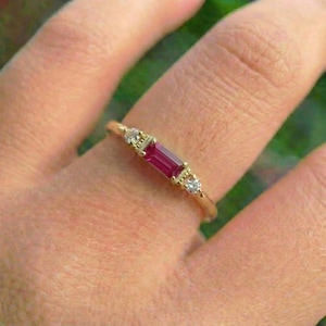 May include: A gold ring featuring a rectangular, deep red gemstone flanked by two smaller, round, clear gemstones. The ring is set on a finger, showcasing its delicate design and elegant style.