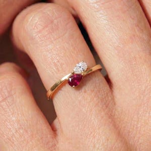 Pode incluir: Um anel de ouro com duas pedras lapidadas em forma de pera num estilo bypass. Uma pedra é uma gema transparente e incolor, e a outra é uma pedra preciosa de cor vermelho-profundo. A banda é feita de ouro fino e polido.