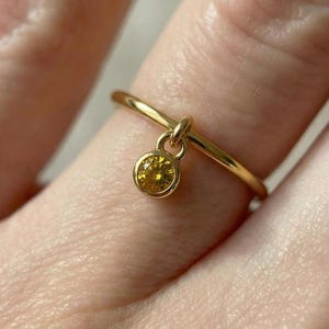May include: A gold ring with a small, round, yellow gemstone pendant. The ring is thin and delicate, with a simple band. The gemstone is a bright, warm yellow color, and is set in a gold bezel.