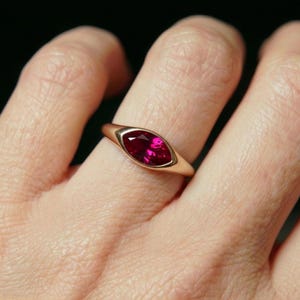May include: A gold ring with a marquise-cut, deep red gemstone. The ring's band is smooth, polished gold, and the gemstone is set in a bezel setting. The ring is shown on a finger against a dark background.