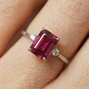 Puede incluir: Un anillo de plata con una gran gema rectangular de color rojo intenso como pieza central. Dos gemas claras, redondas y más pequeñas flanquean la piedra principal. El anillo se encuentra sobre un fondo borroso.