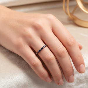 May include: A gold-toned ring with a band of small, rectangular, dark blue stones. The ring is set with small, round gold-colored accents between the blue stones. The ring is on a person's finger.