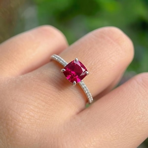 May include: A silver ring featuring a vibrant, square-cut ruby gemstone. The band is adorned with small, sparkling accent stones. The ring is held up against a blurred green background.