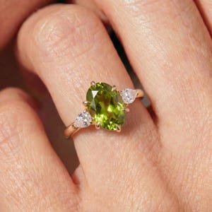 May include: A ring featuring a large oval-cut olive green peridot gemstone set in a polished rose gold band. The central peridot is flanked by two smaller pear-cut clear white diamonds, each secured by metal prongs on a slender gold shank.