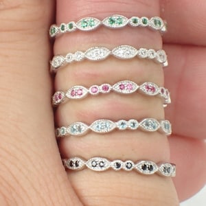 May include: Five delicate silver rings with different colored gemstones. Each ring features a row of small, oval-shaped gemstones set in a silver band. The gemstones are green, white, pink, blue, and black.